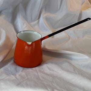 Könnte beinhalten: Eine orangefarbene türkische Kaffeekanne aus Emaille mit einem langen schwarzen Griff. Der Innenraum ist weiß, und die Kanne hat eine klassische, abgerundete Form. Der Griff ist an der Seite der Kanne befestigt. Die Kanne steht auf einer weißen Oberfläche.