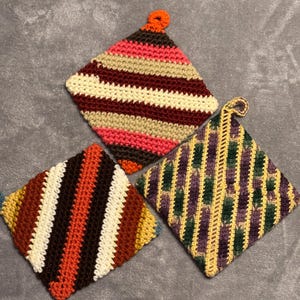May include: Three handmade crochet pot holders in a variety of colors. One is striped with brown, orange, white, and yellow. Another has stripes of pink, brown, beige, and red. The third is purple, green, and yellow.