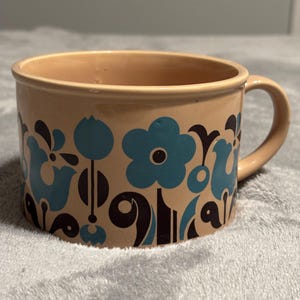 May include: A beige ceramic mug with a handle, featuring a repeating floral pattern in teal and black. The mug has a wide opening and a simple, rounded design, suitable for beverages.