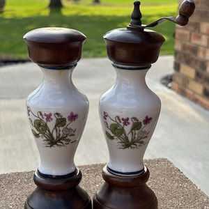 May include: A salt and pepper mill set featuring white ceramic bodies with floral illustrations of purple violets and green leaves. Both pieces have dark brown wooden bases and matching dark brown wooden tops, with a metal crank on the pepper mill.