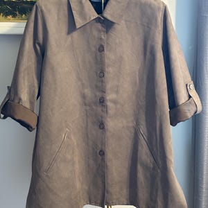 May include: A taupe-colored button-down shirt with short sleeves and a collared neckline. The shirt features a tailored fit with two front pockets and buttoned sleeve cuffs. The fabric appears to have a suede-like texture.
