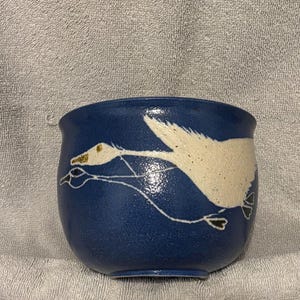 Puede incluir: Un cuenco de cerámica azul con un diseño de ganso blanco. El ganso está representado en vuelo con un pico dorado y está delineado en blanco. El cuenco es redondo y tiene un acabado liso y brillante. El cuenco tiene unos 10 cm de diámetro.