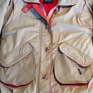 May include: Tan jacket with red trim and accents. The jacket features a collared neckline, a zipper closure, and multiple pockets. The interior lining is red, and the jacket has the brand name "IZZI" on the label.
