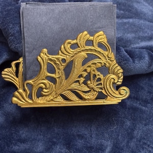 May include: Ornate gold-colored napkin holder with a detailed, swirling design. The holder is filled with dark blue napkins, set against a dark blue velvet background. The design features floral and scrollwork elements.