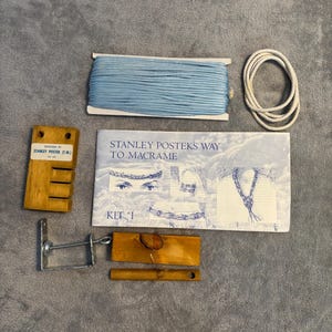 May include: A macrame kit with blue cord, white rope, wooden tools, and a booklet titled "Stanley Postek's Way to Macrame." The kit includes a wooden tool with the text "Designed by Stanley Postek (T.M.)".