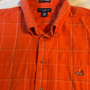 May include: An orange button-down shirt with a plaid pattern. The shirt has a pocket on the left chest and the words "IVY CREW Classics" are printed on the shirt. The shirt is wrinkle resistant.