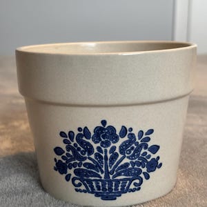 May include: A beige ceramic planter with a blue floral design. The planter has a cylindrical shape with a wide opening and a detailed blue floral pattern on the front. The design includes a basket of flowers.
