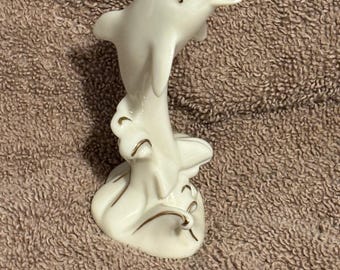 以下が含まれることがあります： 金色のアクセントが付いた白いセラミック製のイルカの置物。イルカは水から飛び出し、体がアーチ状になり、尾が曲がっています。置物は金色のトリムが付いたベースにあります。
