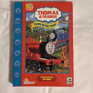 May include: A red and blue Thomas & Friends CD-ROM box featuring illustrations of Thomas the Tank Engine and other characters. The box has the title "Trouble On The Tracks" and is labeled "Interactive CD-ROM". The game is for ages 3 and up.