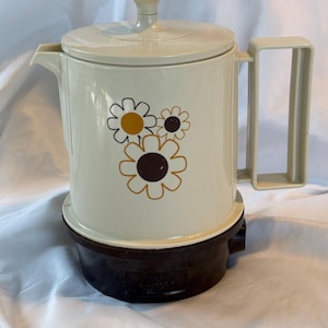 May include: Cream-coloured electric coffee pot with a floral design in brown and yellow. The pot has a lid, handle, and spout. The base is dark brown. The text "POLY HOT POT" is visible on the base.