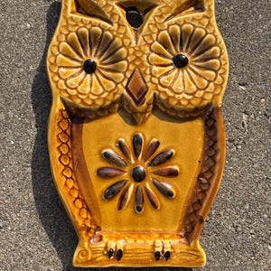 May include: A glazed ceramic wall plaque shaped like an owl in shades of mustard yellow and brown. The owl features large circular eyes, a diamond-shaped beak, and a central flower motif on its chest. A small hole is located at the top center.