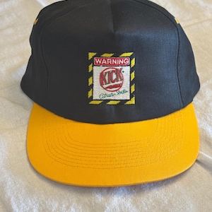 Puede incluir: Gorra de béisbol negra y amarilla con visera plana. La gorra presenta un parche rectangular con la palabra "WARNING" encima de la palabra "KICK" en un círculo rojo, y "Citrus Soda" debajo. La visera y el botón son amarillos.