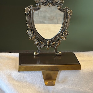 May include: An antique-style Christmas stocking holder with a decorative mirror. The holder is made of bronze-colored metal with floral and scrollwork details. The mirror is shield-shaped and framed by ornate metalwork. The base has a rectangular shelf and a hook for hanging.
