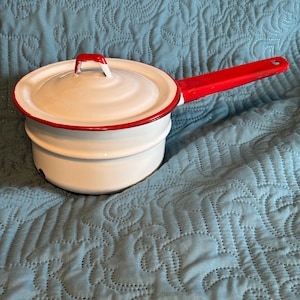 Puede incluir: Un cazo de esmalte blanco con borde rojo y una tapa a juego con asa roja. El cazo tiene un mango largo y recto de color rojo que se extiende desde el lateral. El exterior es de esmalte blanco liso con un borde rojo distintivo arriba.