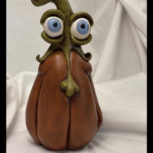 May include: A decorative figurine shaped like a brown pumpkin with a green stem and leaf structure forming a face. The figure features two large, prominent eyes with light blue irises and black pupils, set within green, textured eyelids.