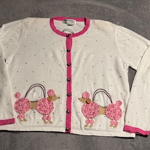 May include: White cardigan sweater with pink trim and black polka dots. Features two appliqued pink poodle designs with brown accents. The sweater has a button-up front and long sleeves.