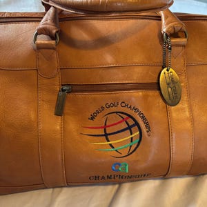 May include: A tan leather duffel bag with a top handle and a zipper closure. The bag features the embroidered text "WORLD GOLF CHAMPIONSHIPS" with a globe design in multiple colors. A metal tag hangs from the side.