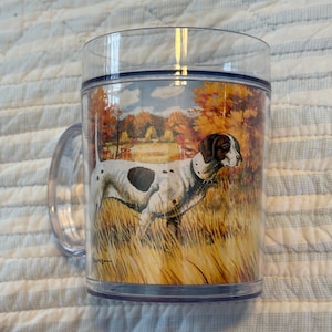 May include: Clear plastic mug featuring a painted image of a Pointer dog in a field with autumn foliage. The dog is white with black spots and a brown head. The handle is clear plastic.