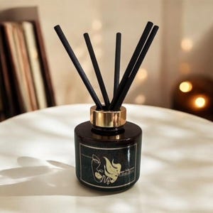 May include: A dark brown glass diffuser bottle with a gold-coloured collar and black reeds. The bottle has a label with a gold-coloured line drawing of a face on a dark green background. The diffuser sits on a white surface.
