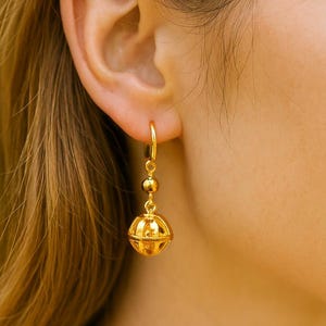 Pode incluir: Brincos de argola dourados com um pendente esférico pendurado. O pendente tem um design em forma de gaiola com uma esfera menor e uma pequena conta acima. Os brincos são mostrados em um fundo desfocado.