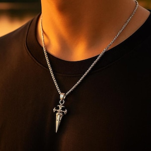 May include: Silver-toned pendant necklace featuring a dagger-shaped charm. The charm has intricate detailing and is suspended from a delicate chain. The necklace is worn against a black shirt, creating a contrast that highlights the jewelry.