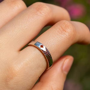 May include: Silver ring featuring a small red gemstone and a geometric pattern. The ring is on a finger, with a blurred backdrop of green and pink foliage.