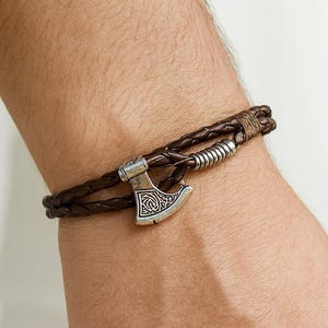 May include: Brown leather bracelet with a braided design and a silver-colored axe charm. The bracelet is wrapped around a wrist, showcasing the accessory's details and craftsmanship. A stylish piece of jewelry.