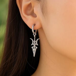 May include: A silver earring with a distinctive, stylized design. The earring has a hoop that connects to a decorative pendant with a symmetrical, arrow-like shape. The pendant has a detailed, openwork design with curved and pointed elements.