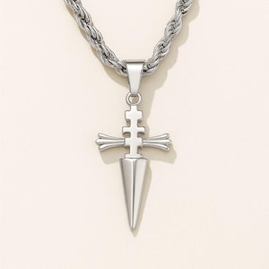 May include: Silver-toned pendant necklace featuring a dagger-shaped pendant with a cross-like design. The pendant hangs from a thick, textured chain. The necklace is displayed against a neutral background.
