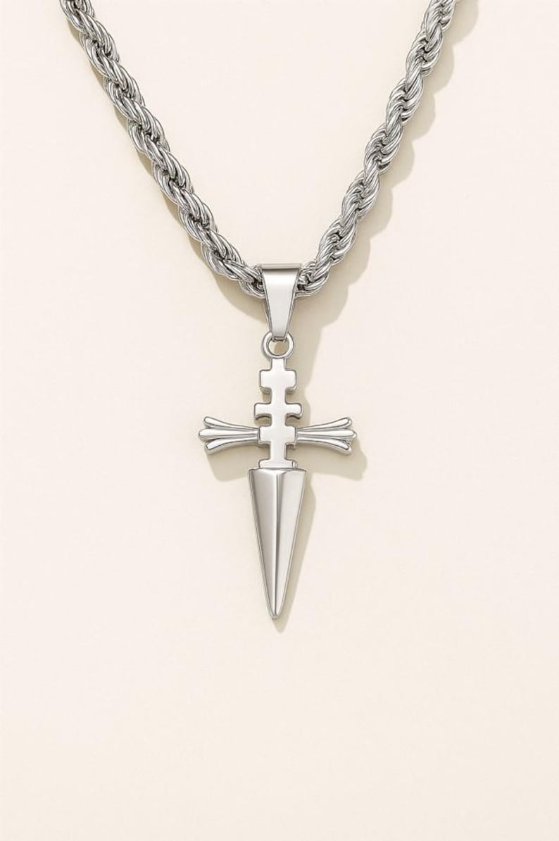 May include: Silver-toned pendant necklace featuring a dagger-shaped pendant with a cross-like design. The pendant hangs from a thick, textured chain. The necklace is displayed against a neutral background.