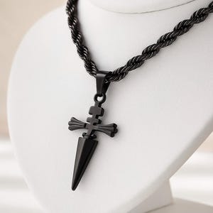 May include: A black pendant necklace featuring a dagger-shaped pendant with a cross-like design. The pendant hangs from a black rope chain. The necklace is displayed on a white bust.