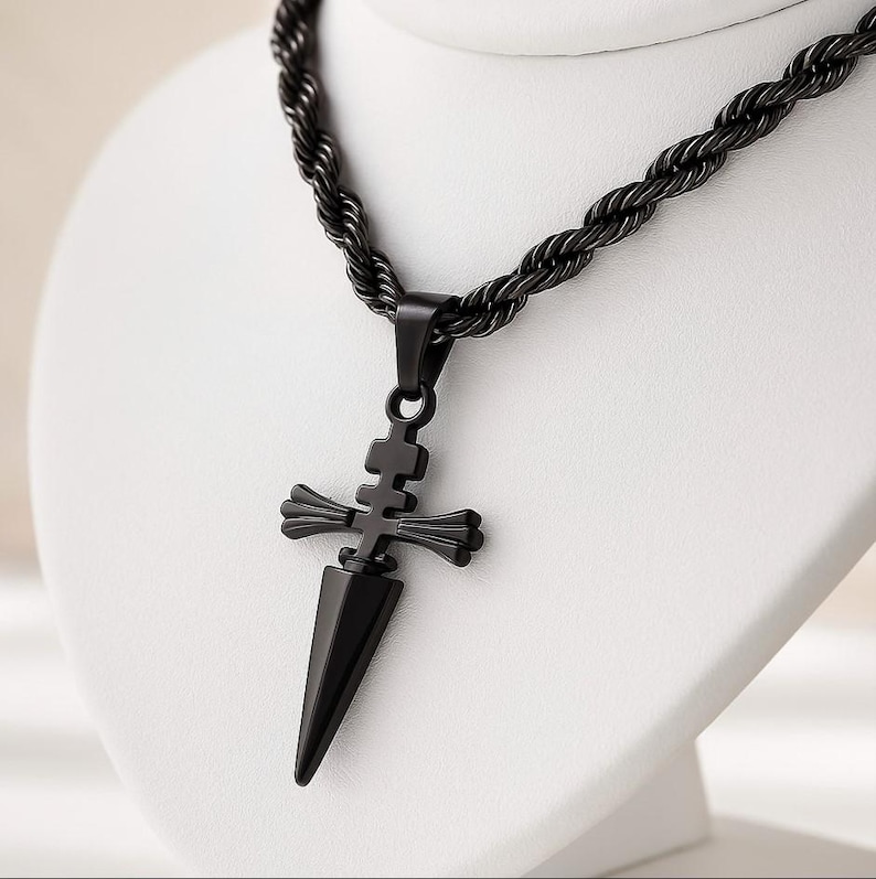 May include: A black pendant necklace featuring a dagger-shaped pendant with a cross-like design. The pendant hangs from a black rope chain. The necklace is displayed on a white bust.