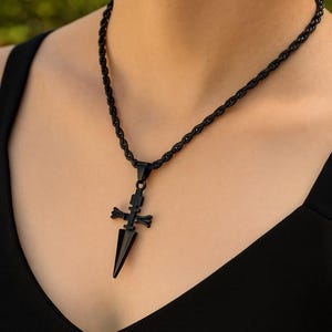 May include: Black necklace with a pendant shaped like a dagger and cross. The necklace has a twisted rope chain. The pendant is a dark, matte black color. The necklace is worn against a black top.