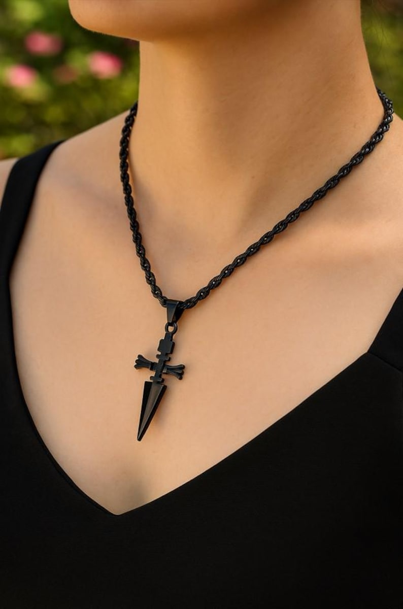 May include: Black necklace with a pendant shaped like a dagger and cross. The necklace has a twisted rope chain. The pendant is a dark, matte black color. The necklace is worn against a black top.