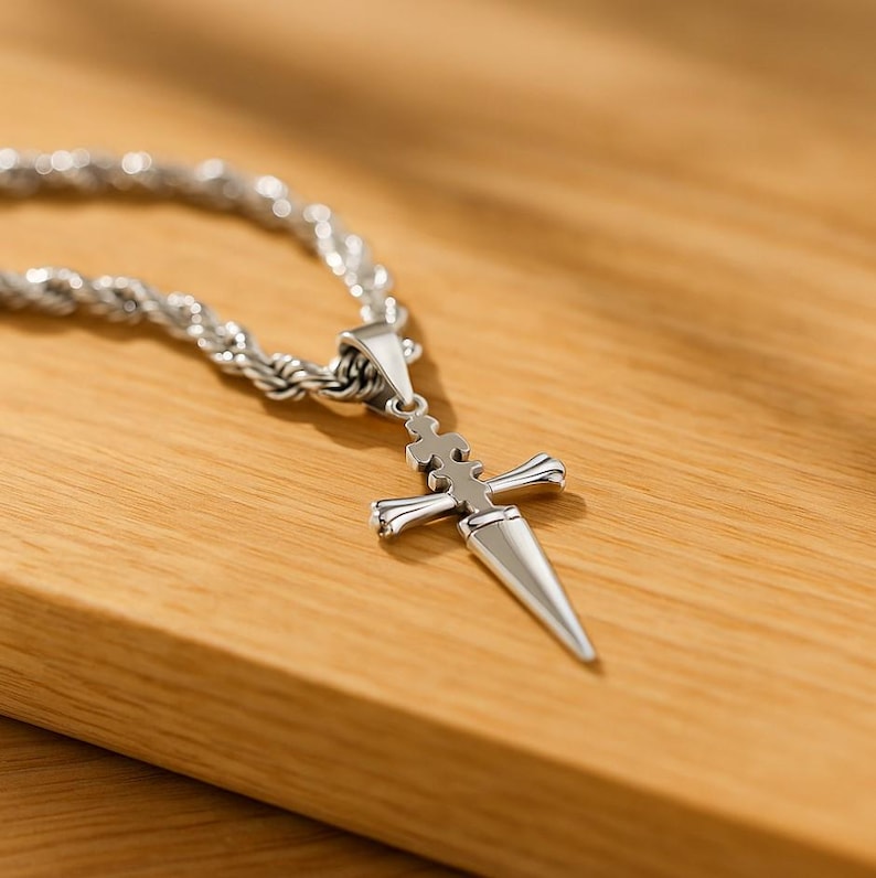 May include: A silver-colored pendant necklace featuring a dagger-shaped charm. The charm has a cross-like design at the top and a pointed blade. The necklace chain is a thick, twisted rope style. The pendant and chain are on a wooden surface.