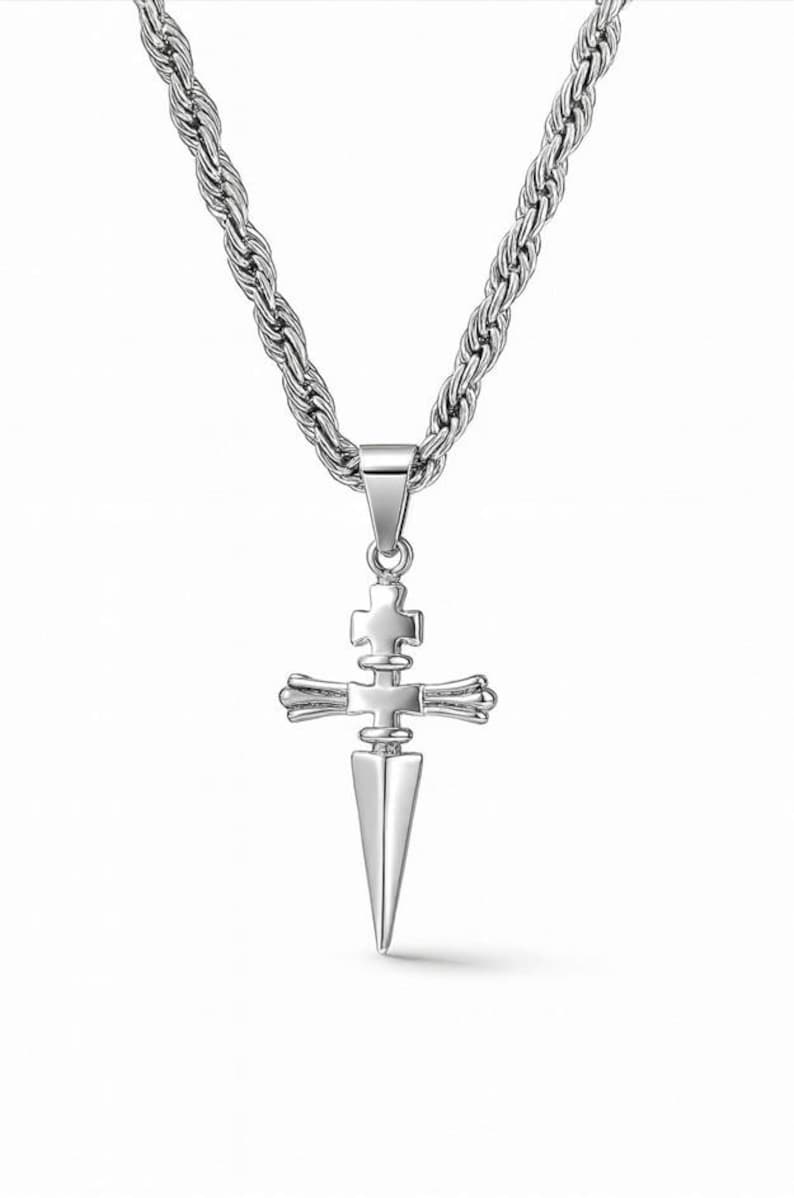 May include: A silver-colored pendant necklace featuring a cross design with a dagger-like point. The cross has a detailed, layered appearance. The chain is a thick, twisted rope style. The jewelry is displayed against a white background.