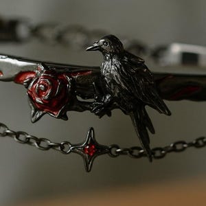 May include: A detailed close-up of a decorative metal object featuring a black raven perched on a branch with a red rose. A chain with a star-shaped charm with a red gem hangs below. The object has a gothic or fantasy theme.