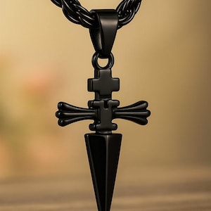 May include: A black pendant necklace featuring a cross-shaped design with a dagger-like point. The pendant hangs from a black, braided chain. The necklace is displayed against a blurred wooden background.