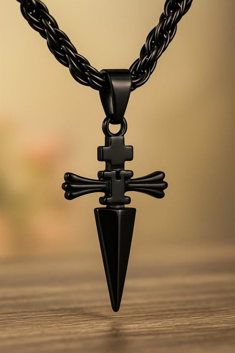 May include: A black pendant necklace featuring a cross-shaped design with a dagger-like point. The pendant hangs from a black, braided chain. The necklace is displayed against a blurred wooden background.