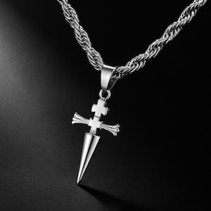 May include: Silver-toned necklace featuring a pendant shaped like a cross with a dagger-like point. The chain is a thick, twisted rope design. The pendant has a cross design at the top and a pointed bottom. The background is black.
