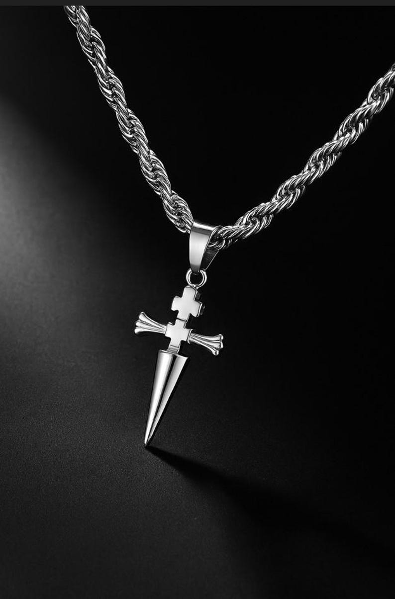 May include: Silver-toned necklace featuring a pendant shaped like a cross with a dagger-like point. The chain is a thick, twisted rope design. The pendant has a cross design at the top and a pointed bottom. The background is black.