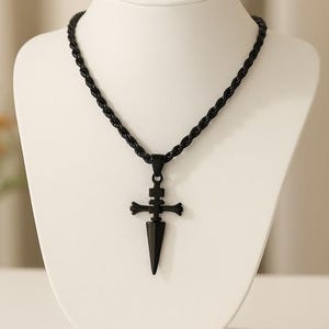 May include: Black pendant necklace featuring a dagger-shaped design with a cross-like embellishment. The pendant hangs from a black rope chain. The necklace is displayed on a white mannequin.
