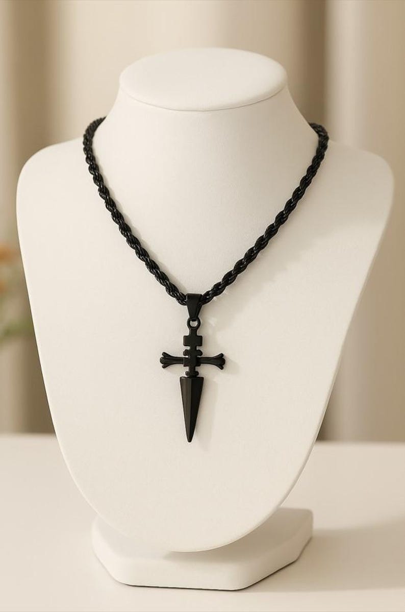 May include: Black pendant necklace featuring a dagger-shaped design with a cross-like embellishment. The pendant hangs from a black rope chain. The necklace is displayed on a white mannequin.