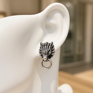 May include: A silver-colored stud earring featuring a wolf's head design with detailed fur and a ring below. The earring is shown on a white background, highlighting the intricate details of the jewelry.