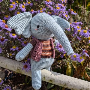 Könnte beinhalten: Eine hellblaue gehäkelte Elefantenpuppe mit großen Ohren und einem Rüssel, die eine rosa-braun gestreifte Weste trägt. Der Elefant sitzt auf einem Holzgeländer, umgeben von lila Blumen.
