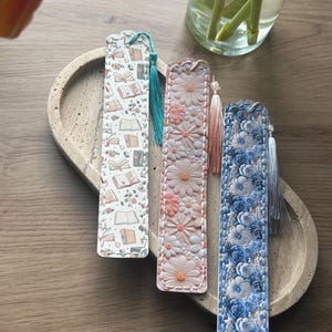 May include: Three decorative bookmarks with tassels. One features a pattern of books and flowers, another has a daisy design, and the third showcases a blue floral pattern. Each bookmark has a different colored tassel.