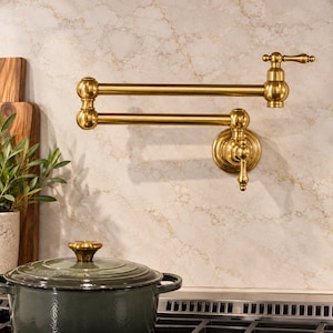 Op de afbeelding: Een aan de muur gemonteerde pannenvul kraan met een geborstelde gouden afwerking, voorzien van een dubbel scharnierende zwenkarm en twee hendels. De kraan is bevestigd op een lichtgekleurde marmeren muur boven een donkergroene gietijzeren pan.