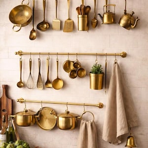 Puede incluir: Tres rieles de cocina horizontales de latón montados en una pared de azulejos, que sostienen utensilios de cocina de latón que incluyen ollas, sartenes, cucharones, espátulas, un batidor, tazas medidoras, una tetera pequeña y dos paños de lino.