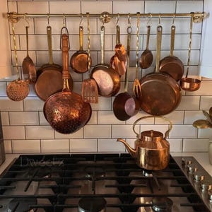 May include: A display of copper kitchenware suspended from a brass rail. The collection features various pots, pans, and utensils, including a colander and a kettle on the gas hob. The kitchen has a white tiled splashback.