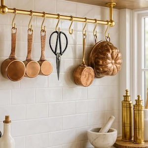 May include: A kitchen scene featuring copper cookware hanging from a brass rod. The items include ladles, a bundt pan, and scissors. Additional brass items and bottles are visible on the counter, creating a warm, inviting aesthetic.